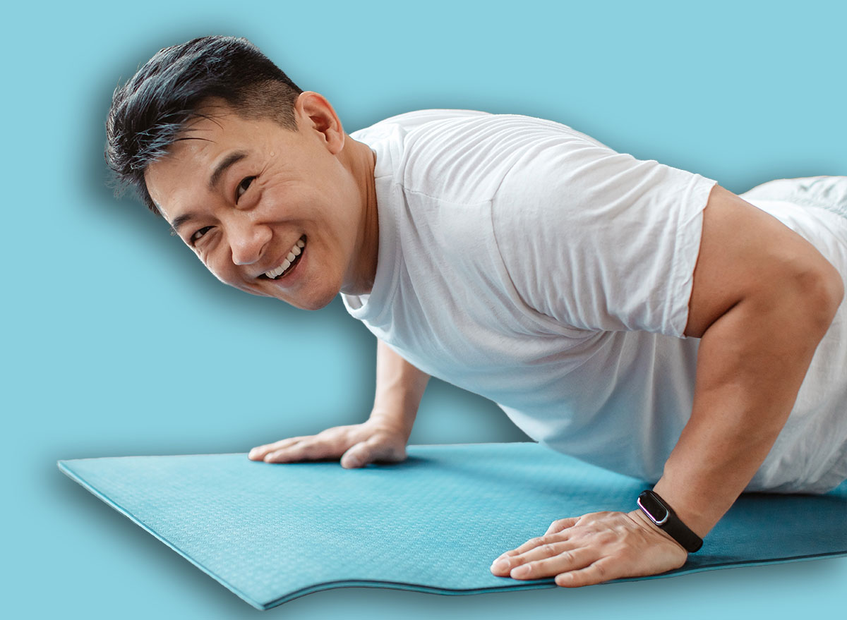 Active asian mature man standing in plank pose or doing push ups on mat at home and smiling at camera. Active korean male leading active lifestyle, exercising indoors