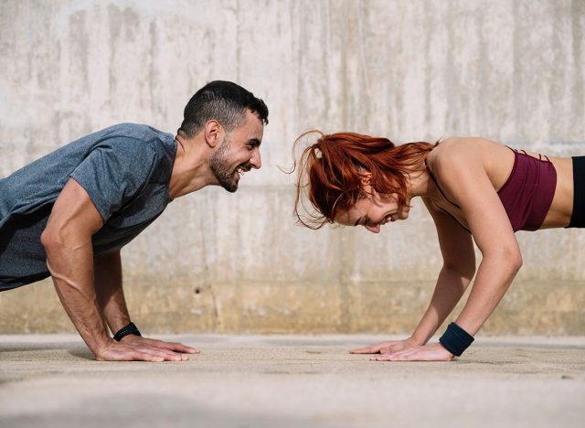 Young adult couple exercising in the floor face to face outside. Joyful athletic male and female doing push ups in the street in sportswear training workout outside.