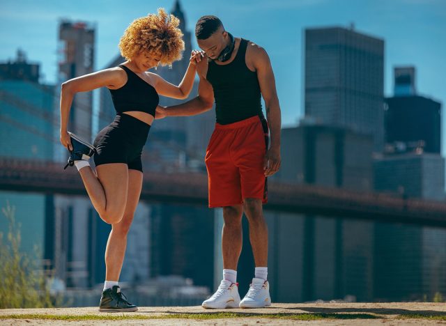 A woman and a man in athletic wear stretching before a workout in an urban setting, with skyscrapers in the background