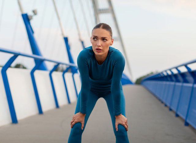 Young tired woman runner in sports outfit resting after running on the bridge in the city in the evening, taking break and breathing