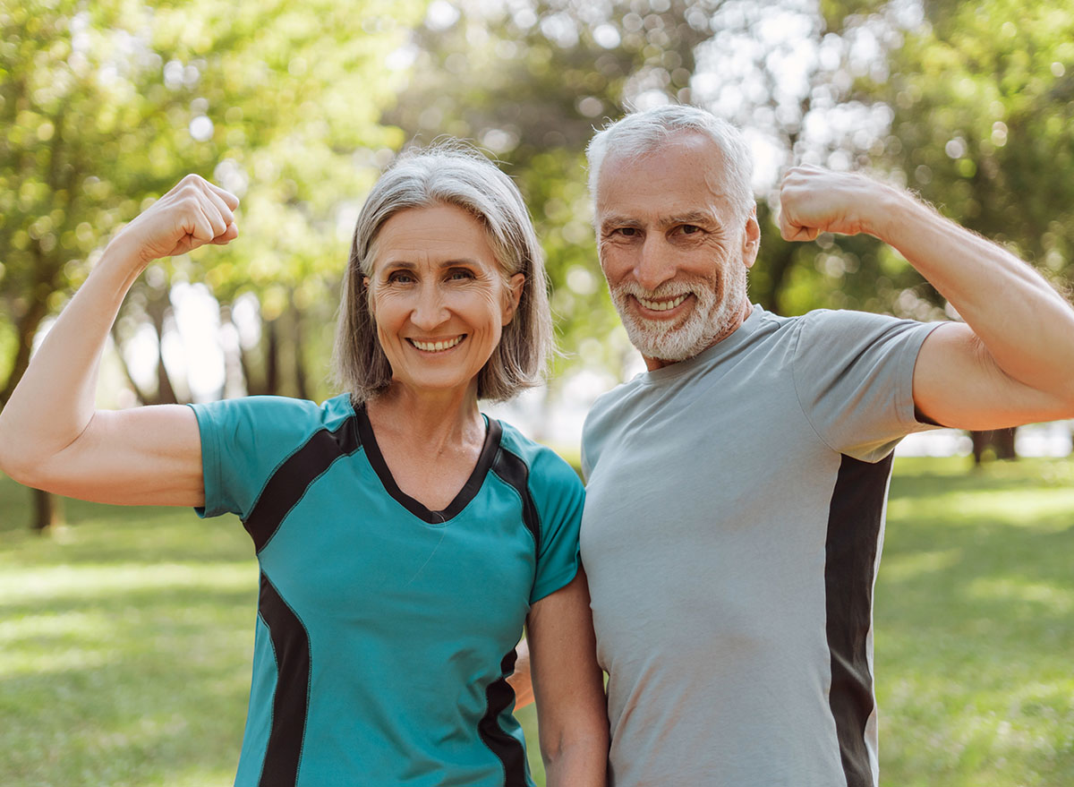 Smiling, attractive senior athletic couple flexing muscles in park, looking at camera outdoors, active lifestyle concept