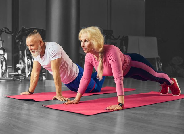 Senior man and woman on sportswear performing push-ups side by side on pink yoga mats in gym. Fitness at any age. Concept of elderly people, sport, active lifestyle, health