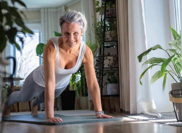 Happy smiling mature middle-aged woman with gray hair does plank exercise. Elderly lady with beautiful athletic body. Anti aging concept
