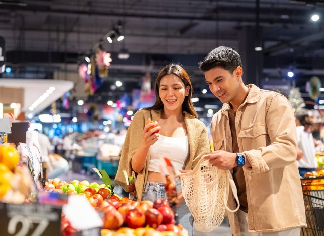 Happy family couple using reusable cotton shopping bag buying food at supermarket. Man and woman enjoy sustainable living and healthy lifestyle choosing organic vegetable and fruit on shelves.