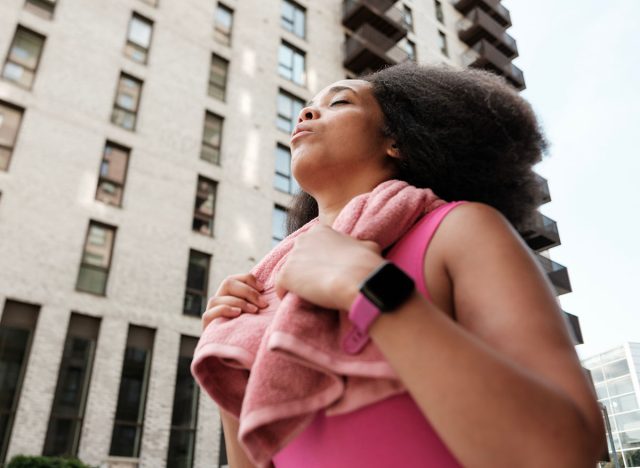 Sporty woman is taking a deep breath of fresh air after her training session, with a pink towel around her neck and a smartwatch on her wrist, standing in front of modern buildings