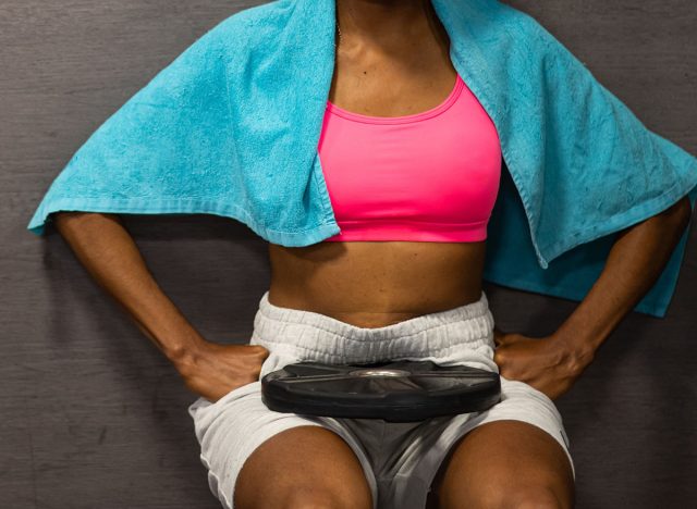 Woman in gym clothes and blue towel around her neck, performing a wall sit exercise with a weight plate resting on her lap, demonstrating strength and commitment to fitness