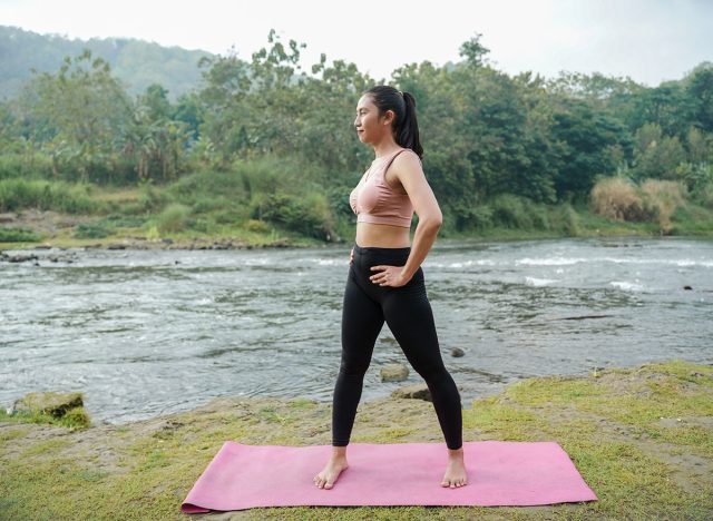 A slender Asian girl with brown skin in sportswear doing stretching movements and rotating her body in the open air on the edge of a beautiful river, before doing sports.