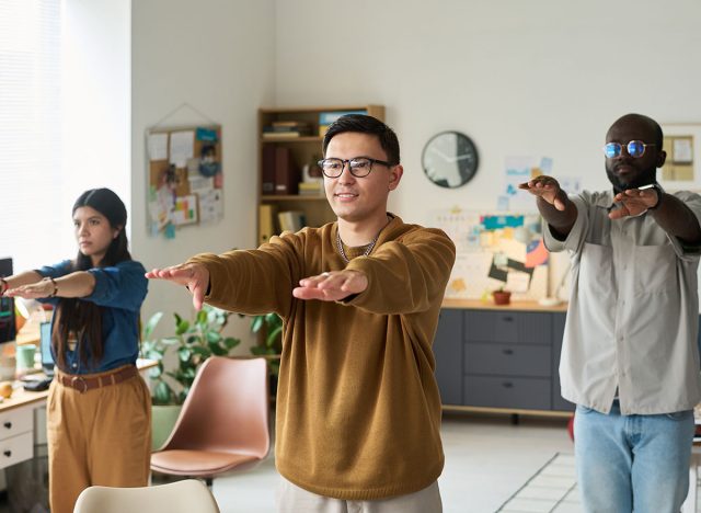 Diverse group of adults standing in office performing stretching exercises with arms extended forward, Asian man, Black man, and woman participating in workplace wellness activity
