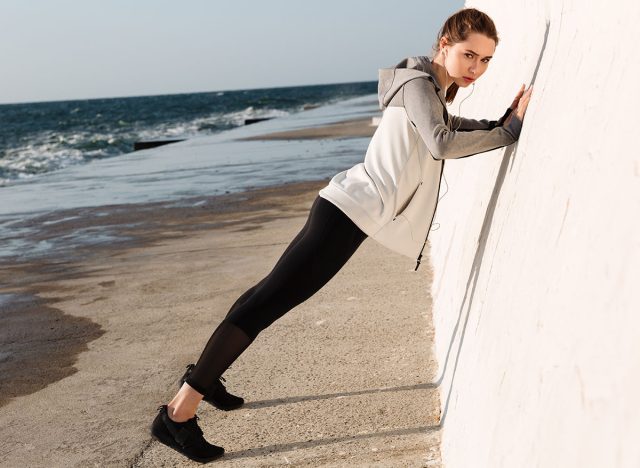 Full-length photo of fit girl doing push-ups while standing near white wall, looking at camera, seaside outdoor