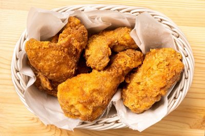 Crispy fried chicken pieces in a white woven basket on light wood. Top view.