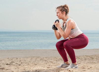 Young attractive woman dressed in pink leggings doing a squat with a kettle bell by the seaside. Fitness woman working out at the beach. Fitness Concept.
