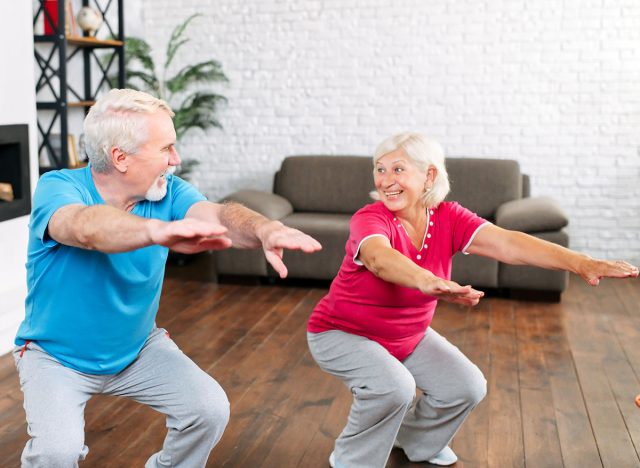 Elderly couple doing squats together at home. Cheerful grey-haired spouses doing exercises, look to each other and smiles