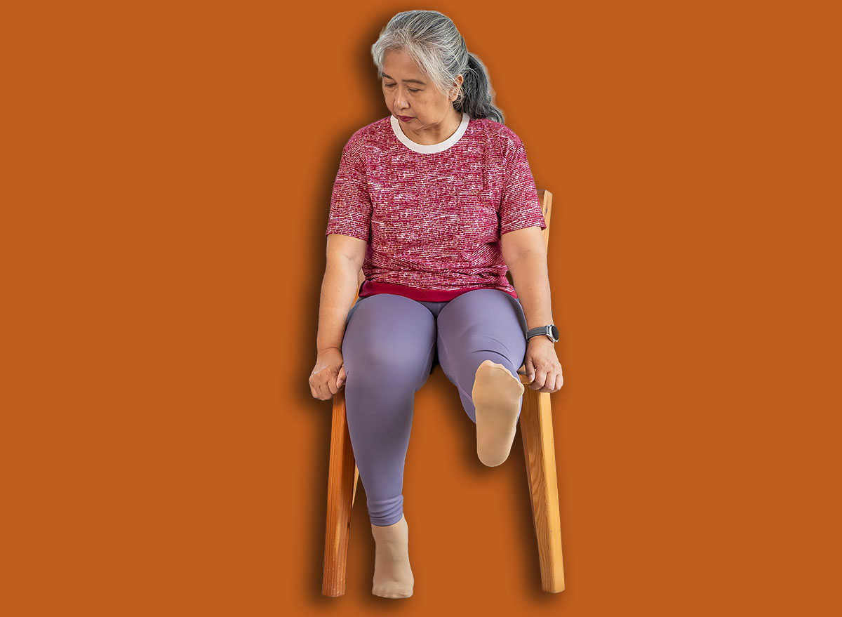 Senior woman sitting on a wooden chair, raising leg to stretch muscles and knees, Training exercise online with tablet In Living Room During Quarantine