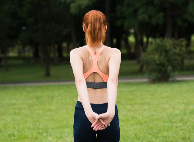 Attractive redhead young woman in sportswear stretching her shoulders in summer in green park. Back view. The girl is warming up. Concept of sport, healthy lifestyle and exercise.