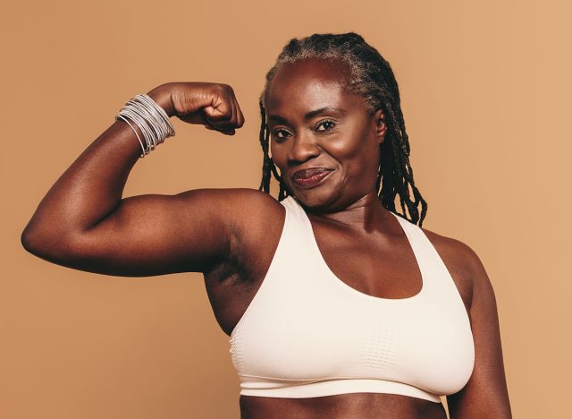 Woman with dreadlocks looking at the camera while flexing her bicep. Mature woman standing against a brown background in sports clothing. Sporty black woman maintaining a fit lifestyle.