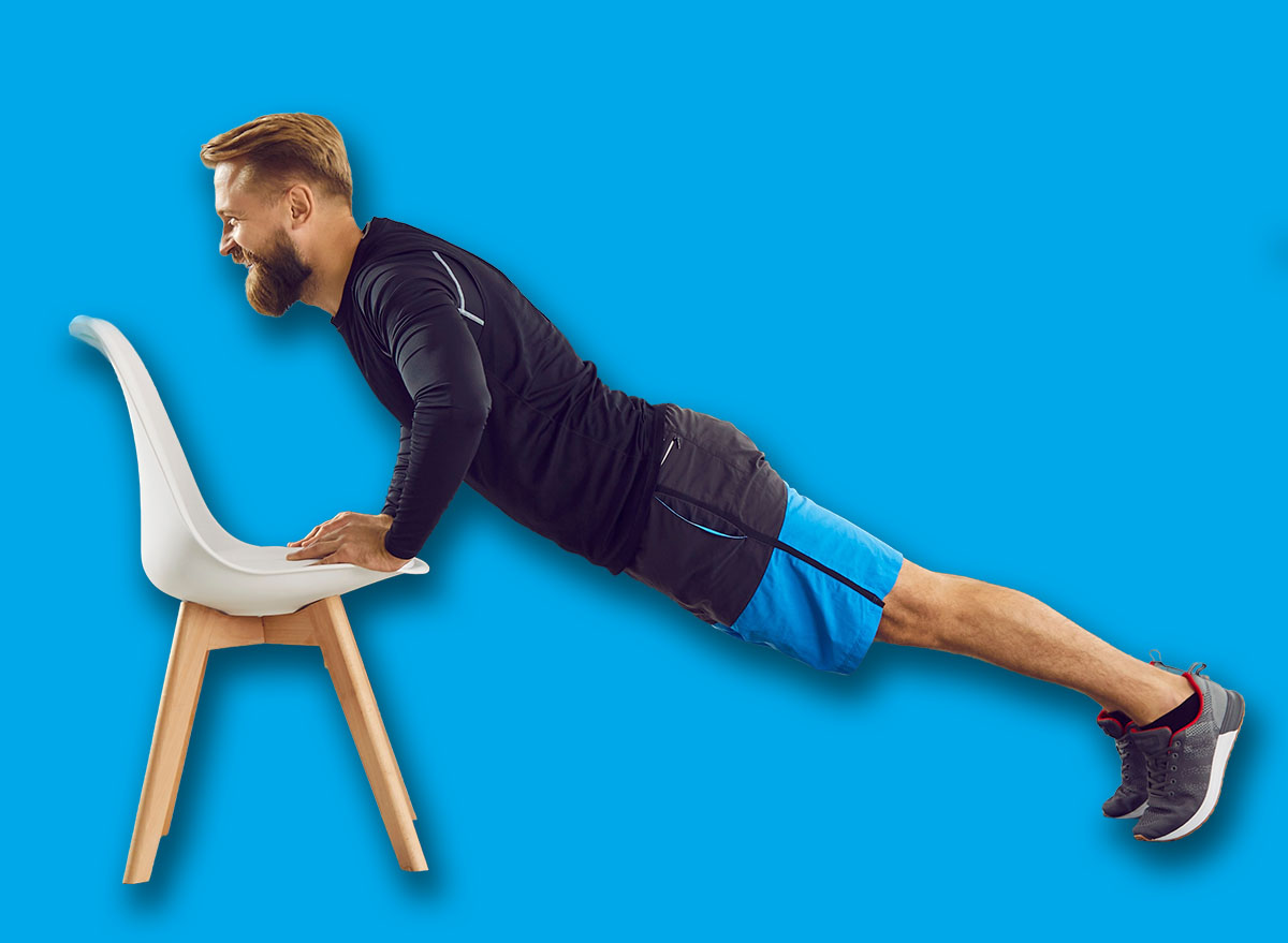 Gym at home. Athletic man is doing push up exercise with chair at home in his spacious living room with modern interior. Side view of motivated millennial guy training his muscles by doing sports.