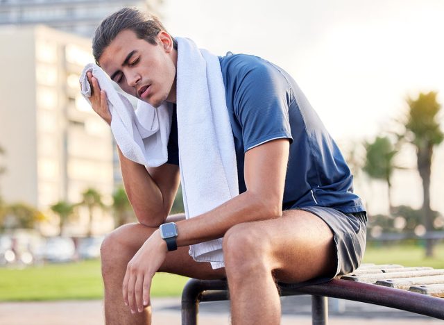 Fitness, exercise and tired man with a towel on park bench to relax or rest after running for cardio. Sports person with fatigue and sweating outdoor post workout or training for health and wellness