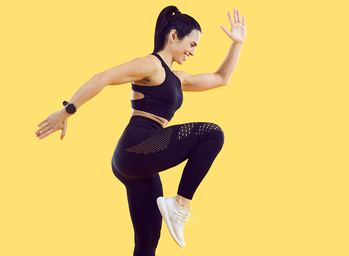 Active sporty woman doing high knees exercise isolated on vivid yellow background. Full length side view of smiling woman with beautiful athletic body dressed in black sportswear doing sports workout.