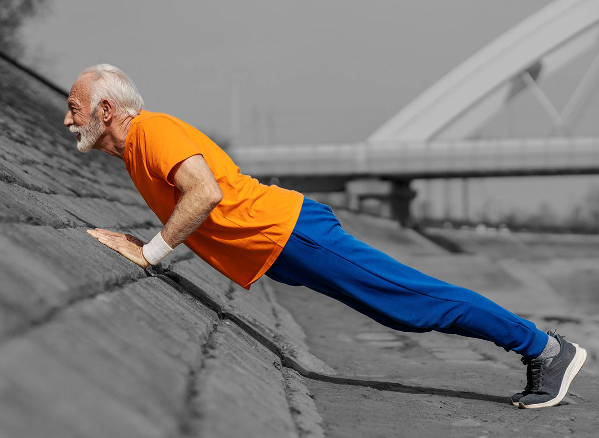 Profile view of an old athlete doing pushups and strength exercises on river shore.