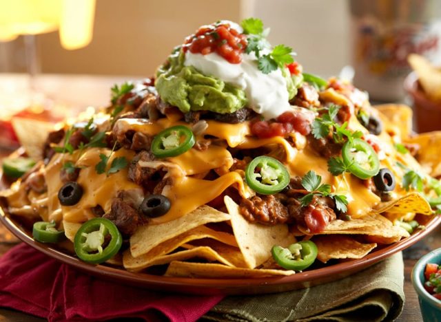 Loaded nachos piled high with melted cheese, jalapeños, black olives, salsa, sour cream, and guacamole, served on a party platter.