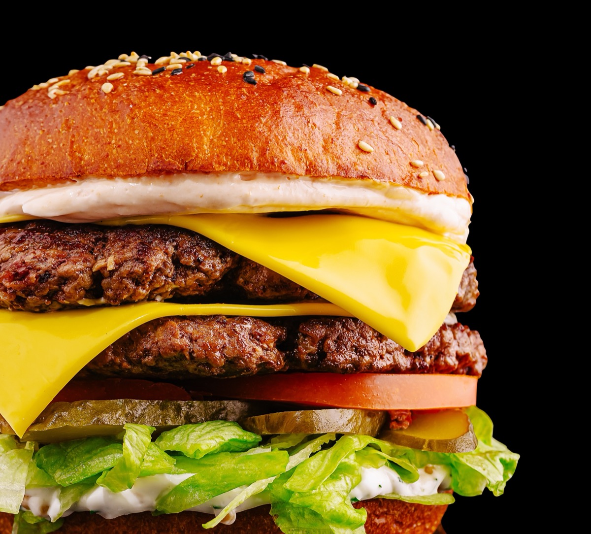A mouthwatering burger features two juicy beef patties layered with cheddar cheese, fresh lettuce, sliced tomatoes, pickles, and creamy sauce, all in a toasted sesame seed bun.