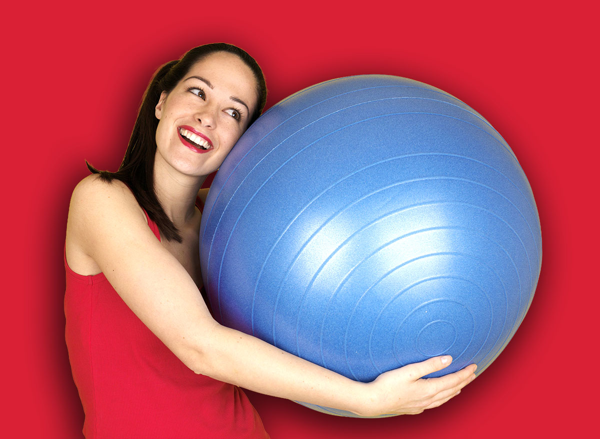 Happy Fit Confident Sporty Young Woman Holding A Blue Swiss Exercise Ball Smiling Isolated On Yellow Alone Wearing Red Sports Top