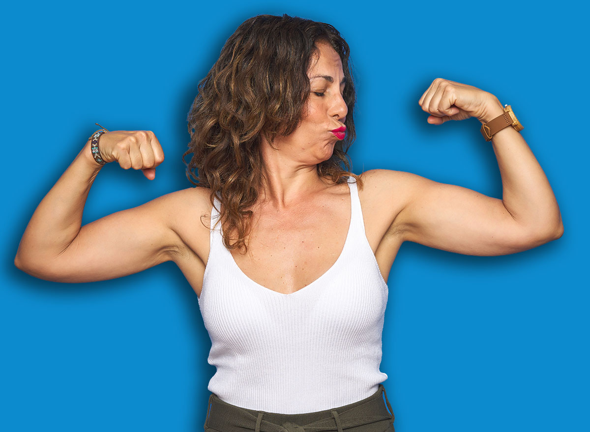 Middle age senior woman with curly hair standing over blue isolated background showing arms muscles smiling proud. Fitness concept.