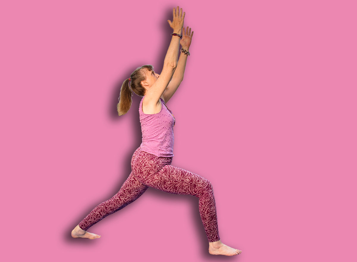 A mature female yoga teacher demonstrates the Warrior I pose in front of a peaceful lake