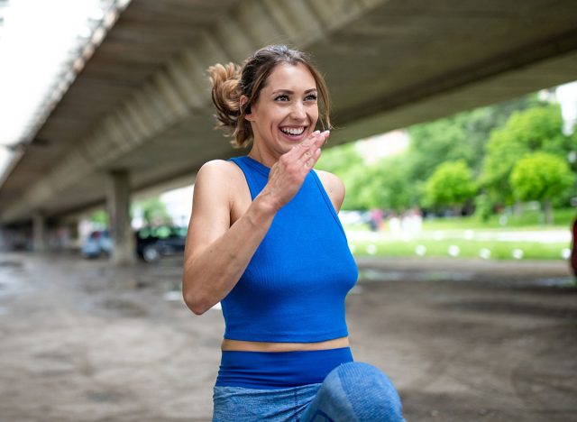 Young woman doing cardio high knee exercise outdoors. Fit athlete smiling exercising under bridge.