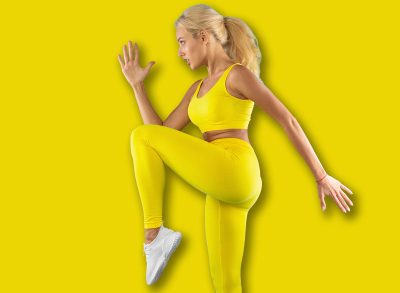 Training Concept. Confident Focused Fit Young Lady In Sportswear Jumping And Lifting Leg Up, Doing High Knee To Elbow Exercise On Blue Studio Wall. Profile Side View Portrait. Energy And Sport