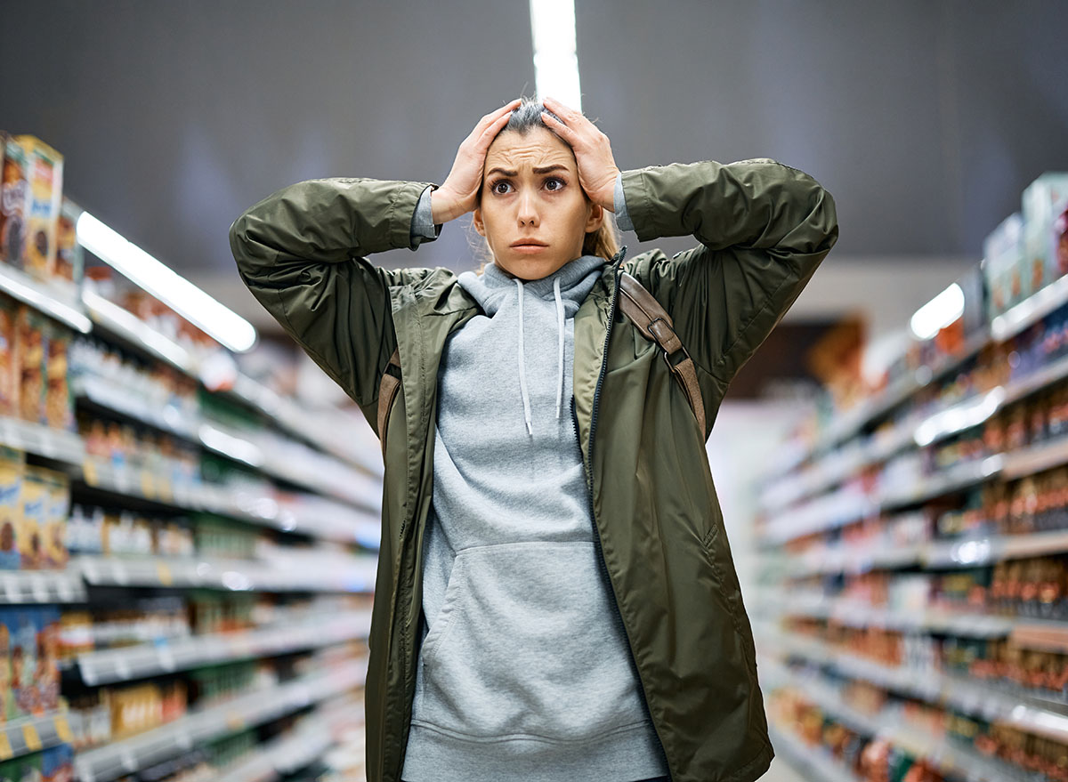 Young woman feeling shocked about rising food prices while standing among the aisle with groceries in supermarket.