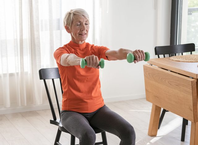 Smiling beautiful senior woman health instructor doing chair exercises with dumbbells