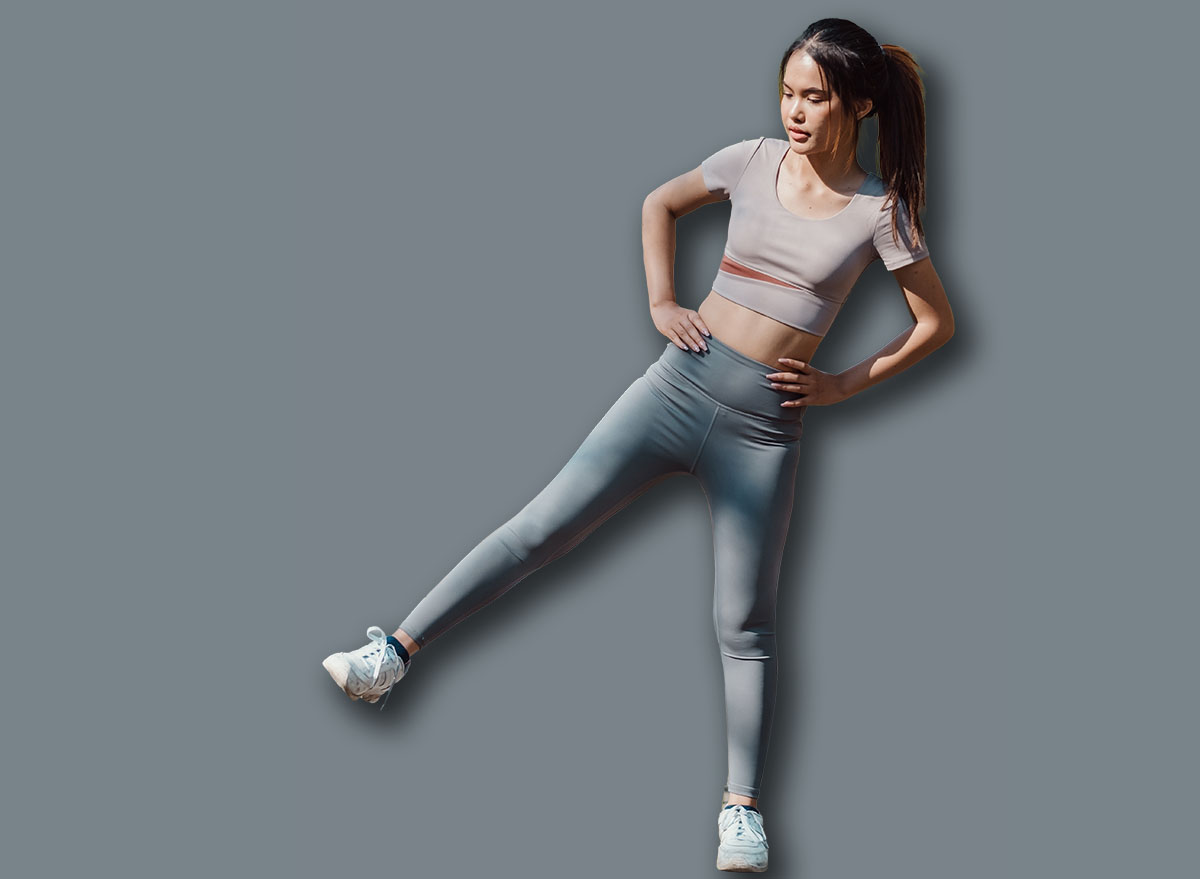 Young woman in athletic wear exercising outdoors, performing a side leg lift on a park path surrounded by trees.