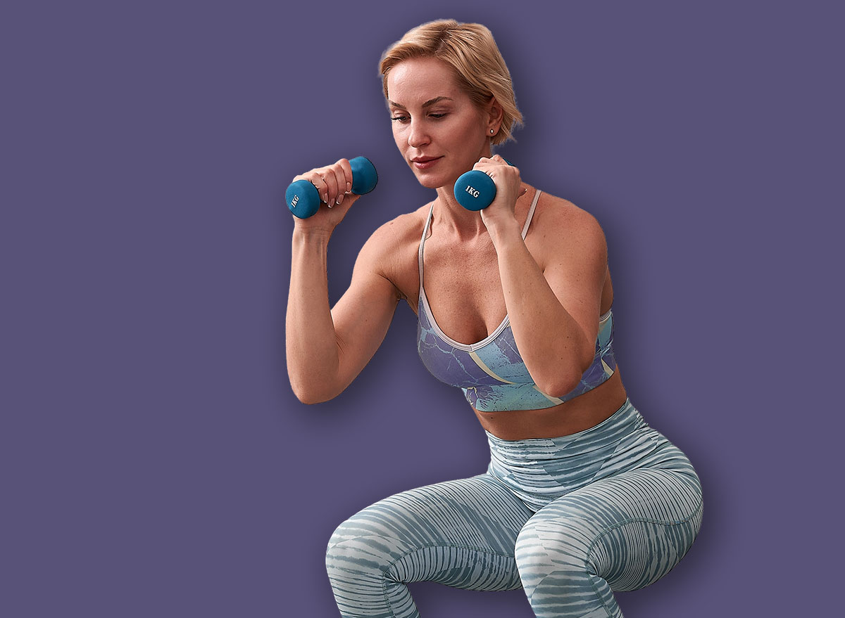 Female trainer performs a squat with dumbbells in hands, arms bent at the elbows and pressed to the chest. Beautiful sporty woman doing power fitness exercise at sport gym.