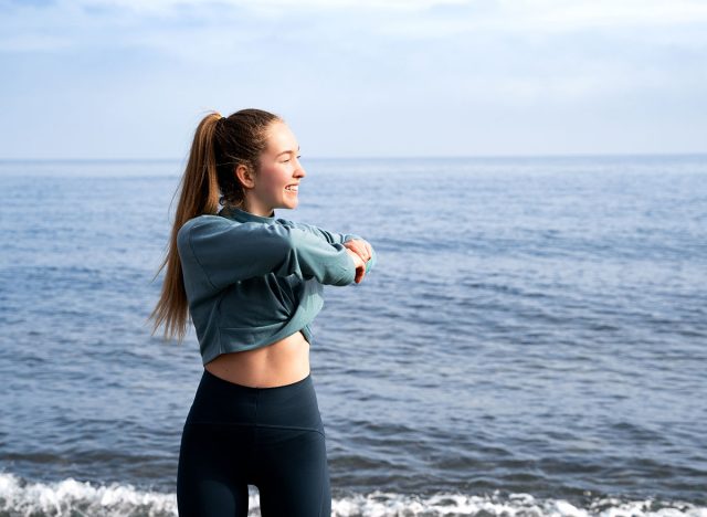 Woman doing body twist stretching sea background. Slim fit girl athlete in sportswear warming up before running, standing on the beach. Sport exercise outdoors. Morning workout in nature, wellness