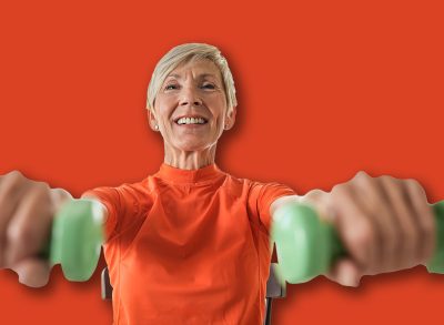 An elderly woman sits on a chair, lifting green dumbbells while smiling. She exercises in a bright living room with plants, promoting strength, fitness, and an active lifestyle.