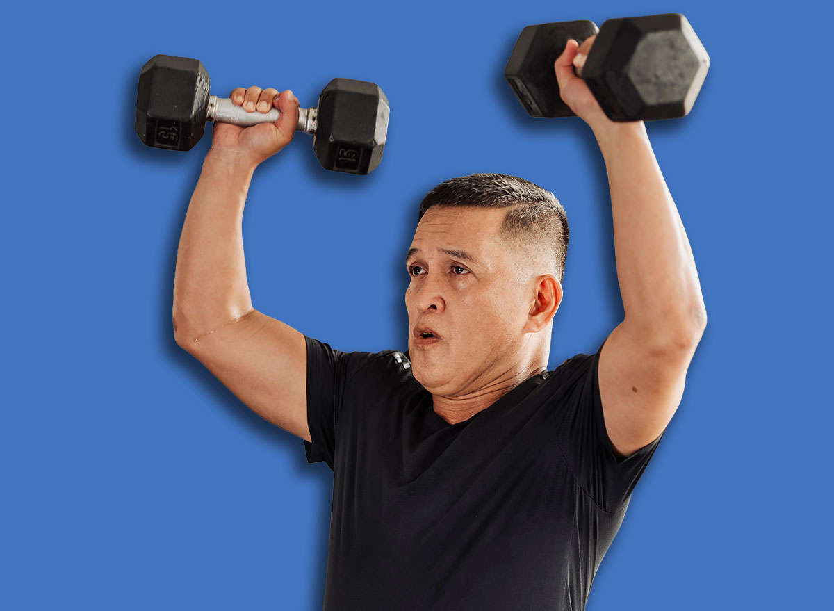 Southeast Asian man in his mid 50s performs an overhead dumbbell shoulder press at an open-air gym with proper form and dedication.