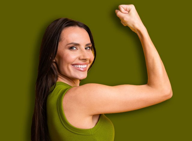 Confident young woman smiling and flexing her arm against a bright yellow background, symbolizing strength, positivity, and cheerful energy.
