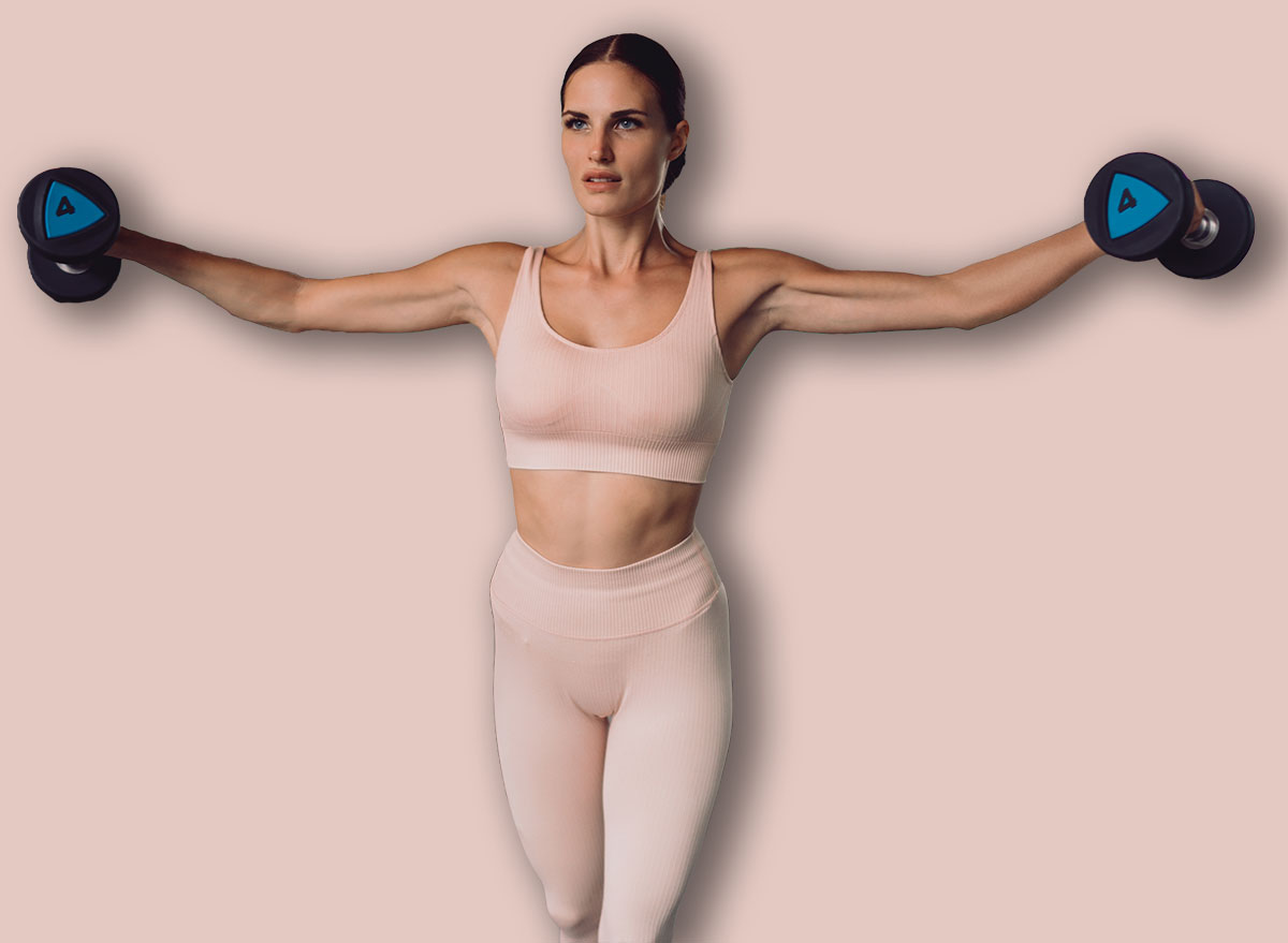 Fit woman holding dumbbells extended outward during lateral raise, showcasing full-body engagement, arm activation, and disciplined strength movement in modern gym.