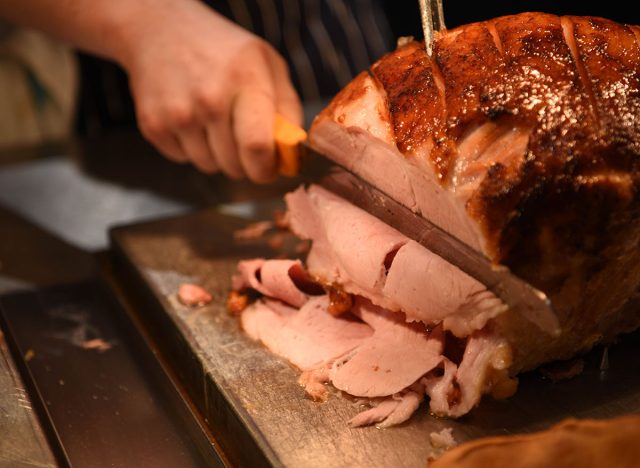 food photography image of roast gammon ham joint of meat being carved with mans hand holding a knife slicing the cooked dinner on a board with blur background and space