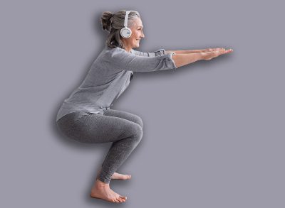 side view of cheerful middle aged woman in sportwear and headphones doing squat on mat at home