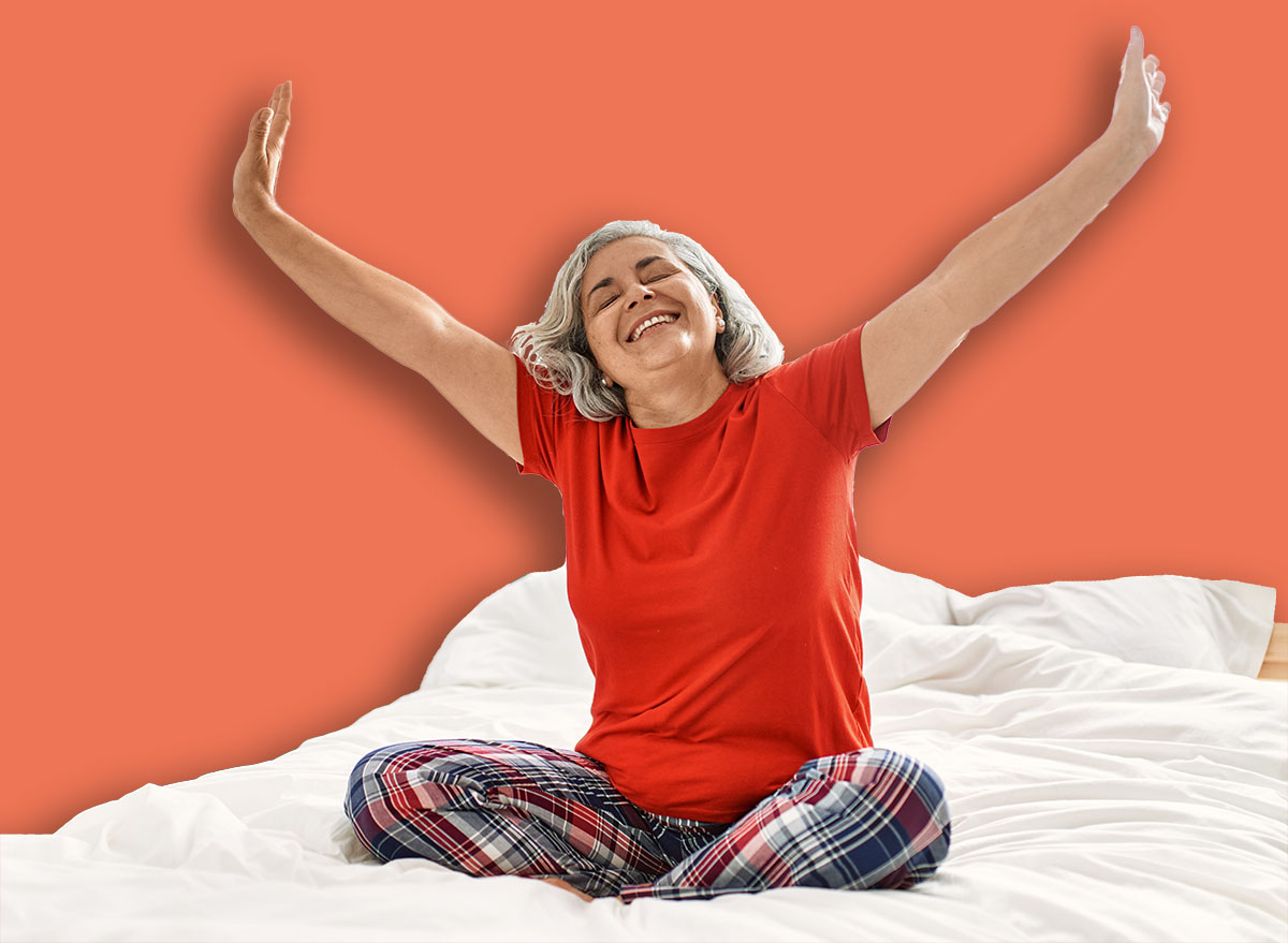 Middle age grey-haired woman smiling happy sitting on the bed with hands raised up at bedroom.