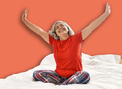 Middle age grey-haired woman smiling happy sitting on the bed with hands raised up at bedroom.