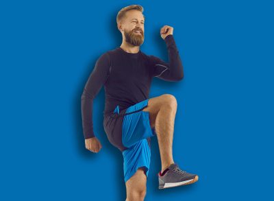 Male athlete in modern activewear having his routine fitness workout at home. Happy handsome bearded athletic man in sports shorts doing high knees walk in place exercise on rubber mat in living room