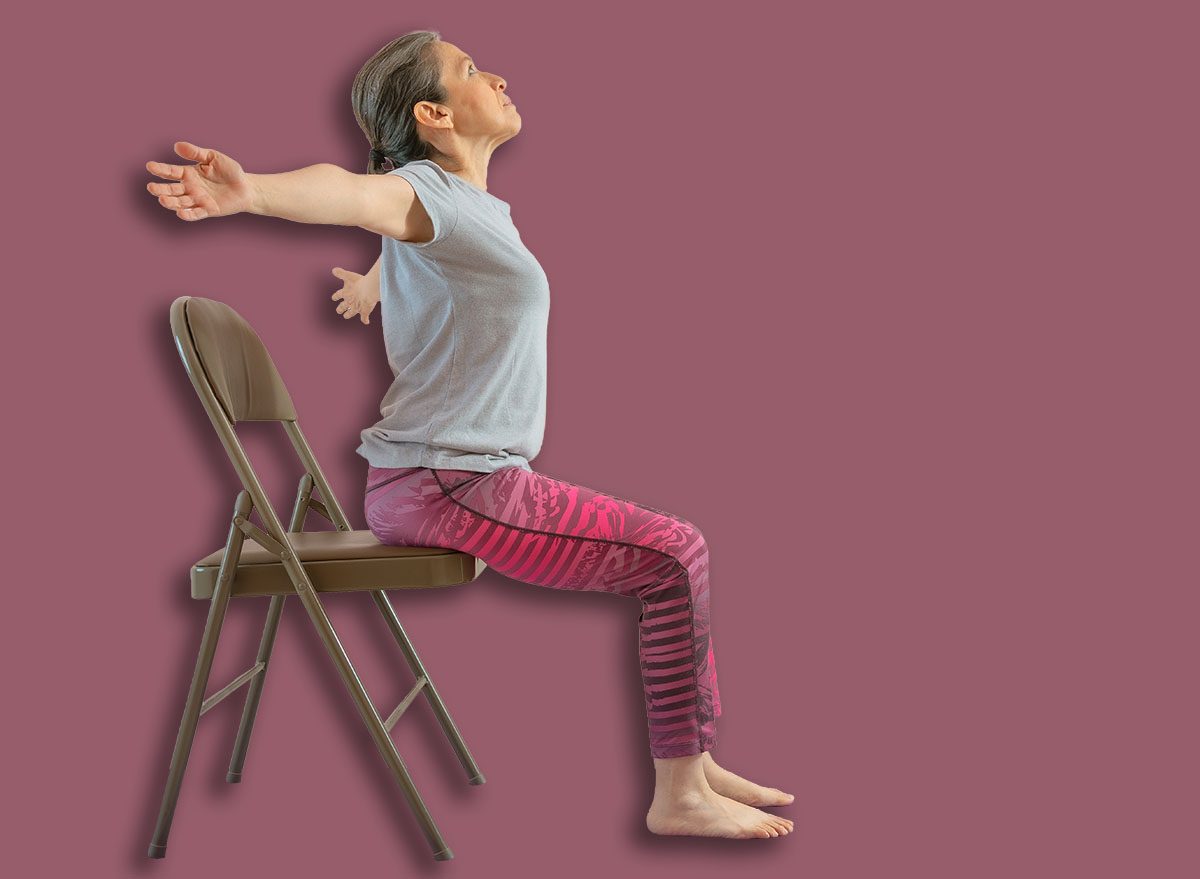 Full length shot of energetic mature woman making exercising and practicing restorative yoga on mat and chair at home because of social distancing, wearing sports clothes.