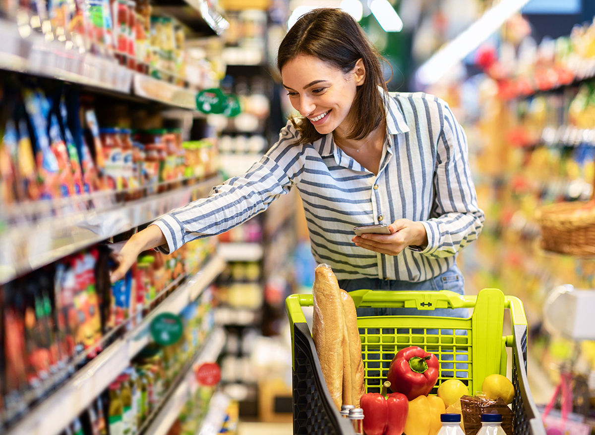 Shopping. Young Smiling Woman Holding And Using Mobile Phone Buying Food Groceries Standing In Supermarket. Female Customer With Smartphone Taking Healthy Products From Shelf At The Shop