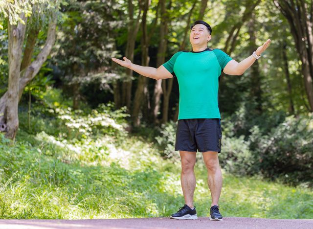 Sporty happy Japanese guy practicing yoga meditation, receives positive energy, healthy lifestyle. Young man standing making breath exercises, relaxing in park on sunny summer day. Active sportsman.