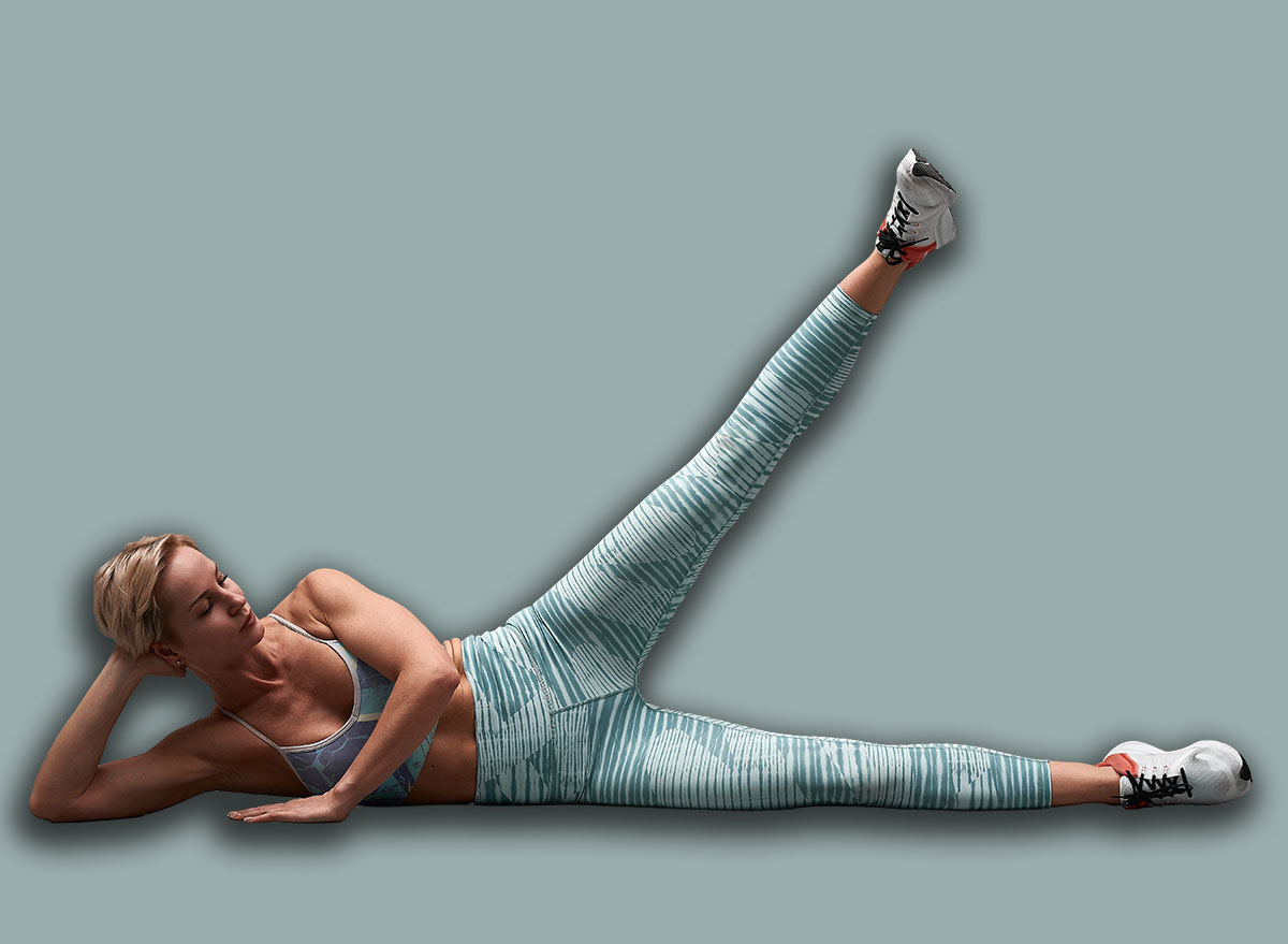 Female trainer lying on her side lifts straight leg up. Pretty girl wearing leggings and short top lying on her side at gym, training body core and balance, strengthening abs muscles.