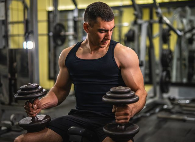 young muscular man hold dumbbells and sit on sports bench in the gym