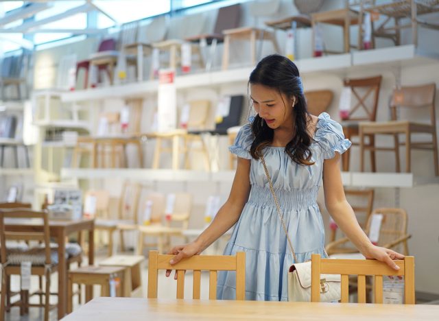 Young asian woman in light blue dress examining wooden chairs at furniture store, interested in home decor and furnishings